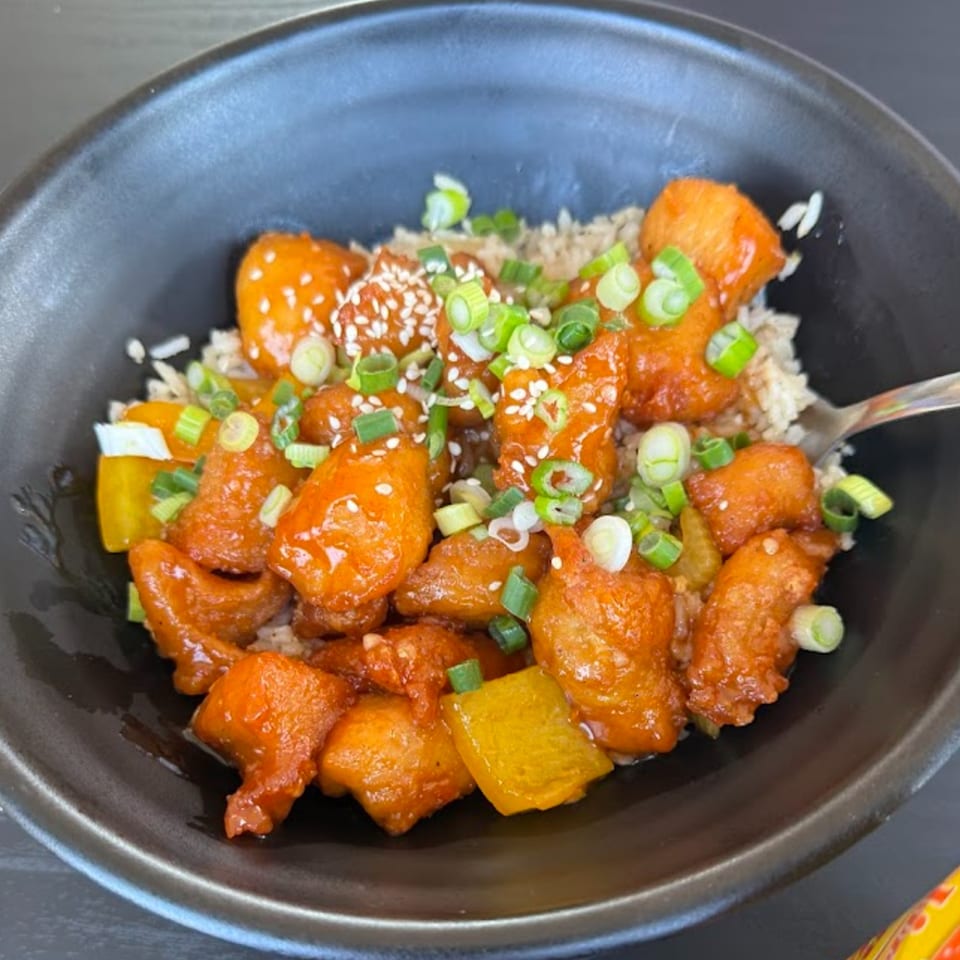 Sweet and sour chicken with rice, bell peppers, sesame seeds, and green onions in a black bowl.