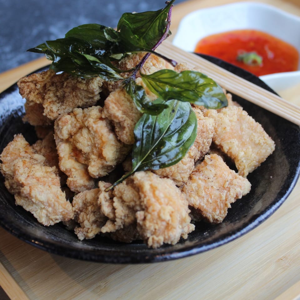 Crispy fried chicken pieces with basil leaves, chopsticks, and a side of red dipping sauce.