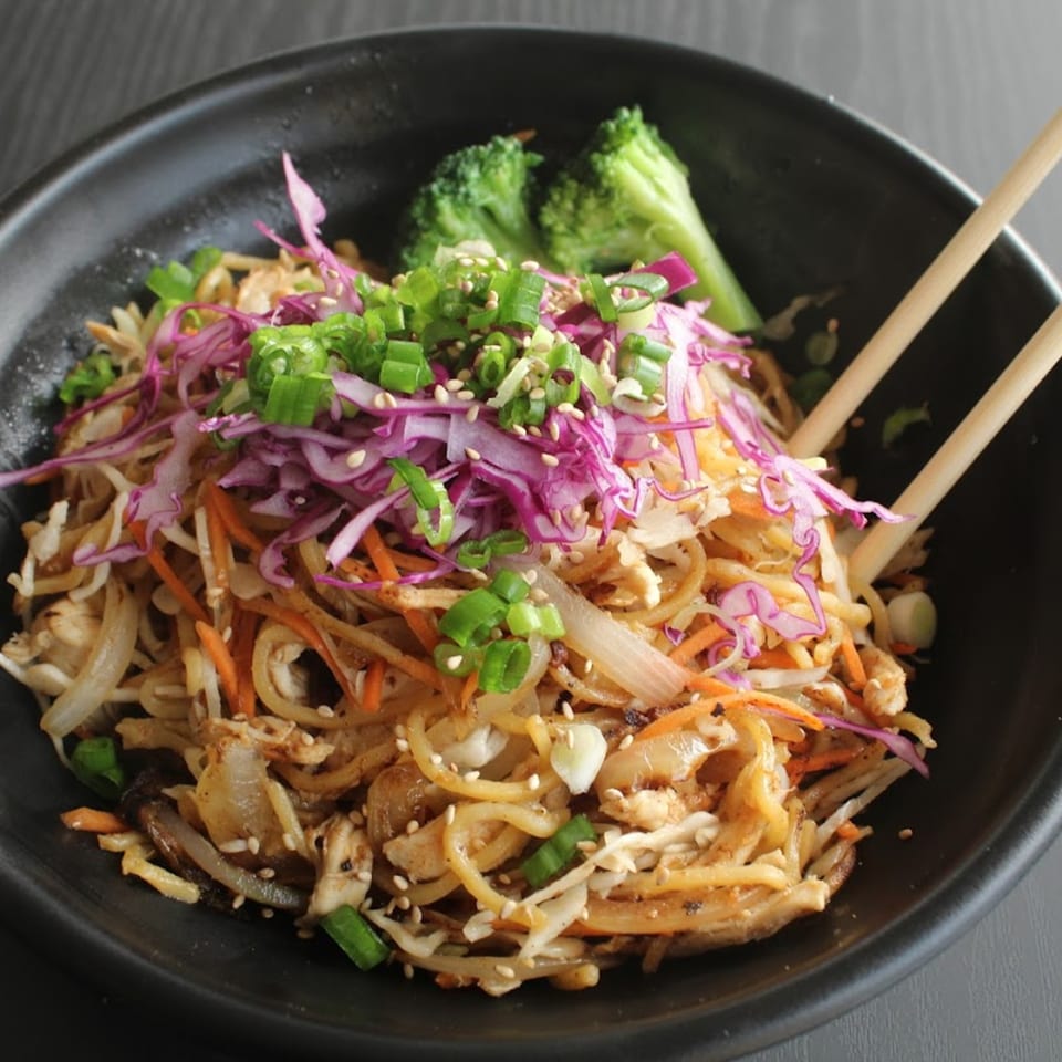 Asian-fusion noodles with chicken, vegetables, sesame seeds, and chopsticks in a black bowl.