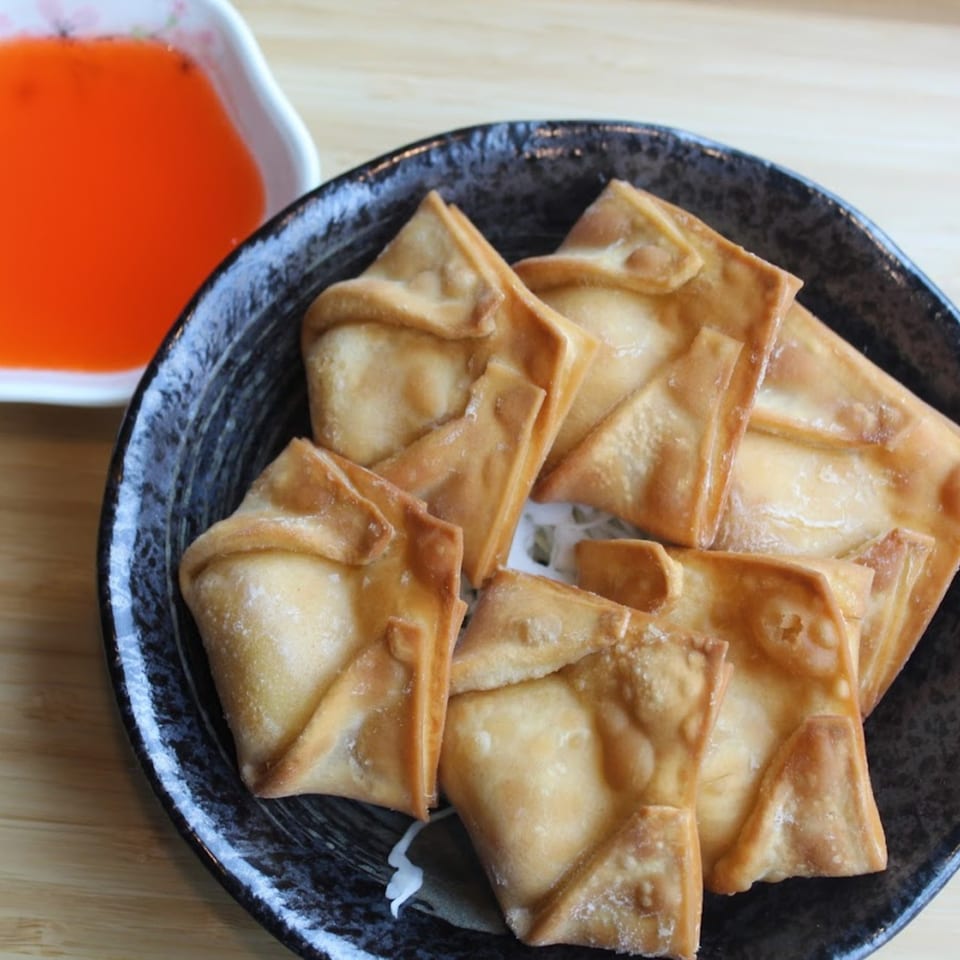 Golden-brown fried wontons on a dark plate, with a side of orange dipping sauce.