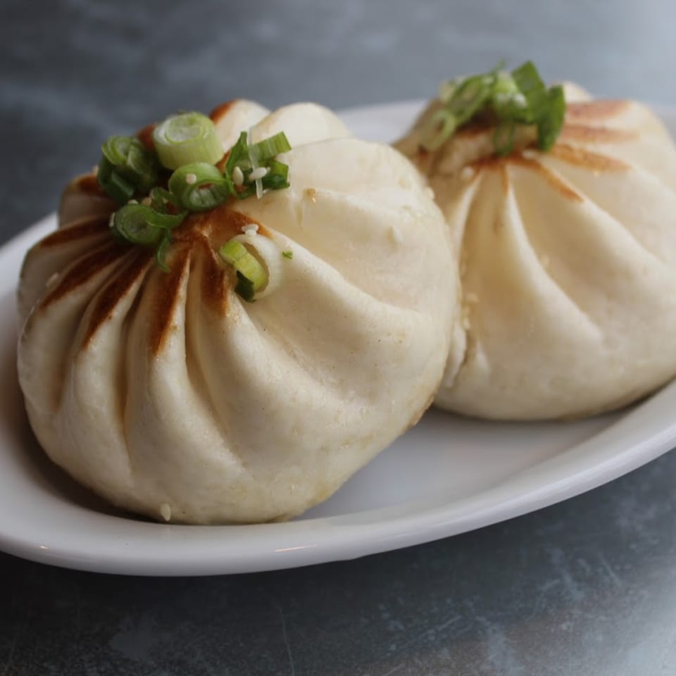 Two steamed buns, garnished with green onions and sesame seeds, on a white plate.
