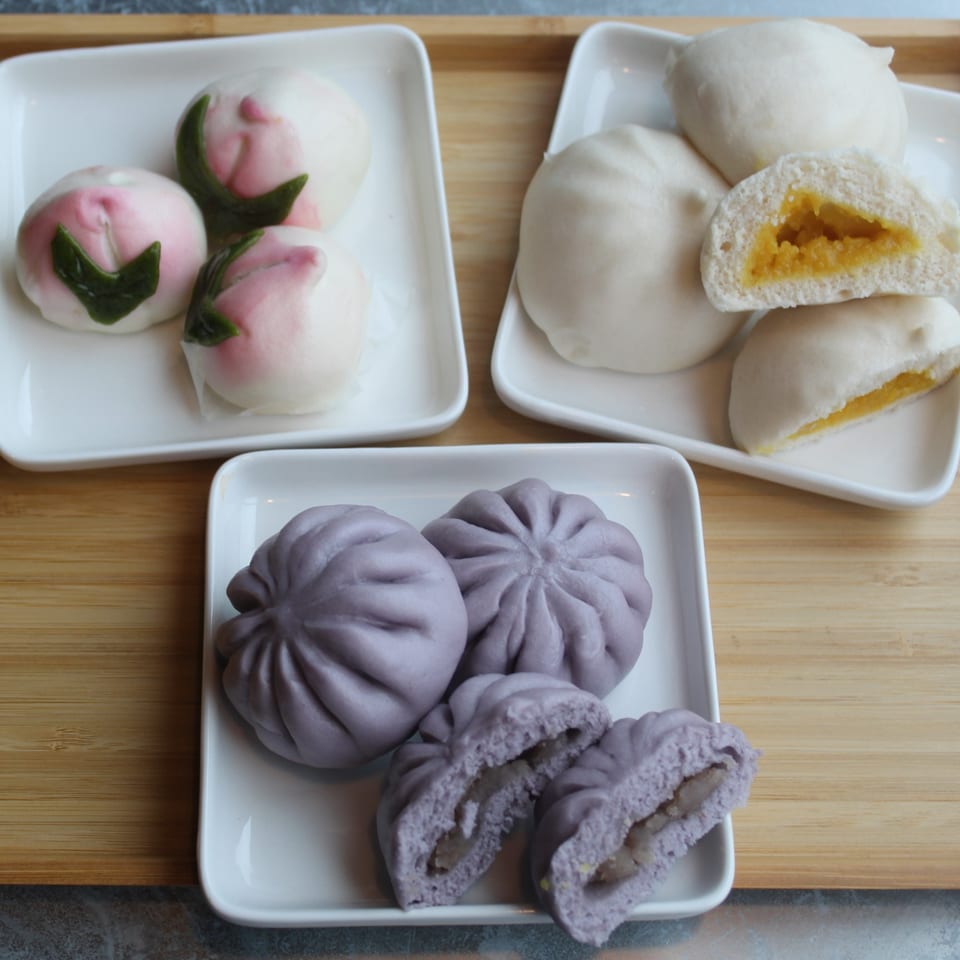 Three plates of colorful steamed buns, including peach-shaped, purple taro, and yellow custard fillings.